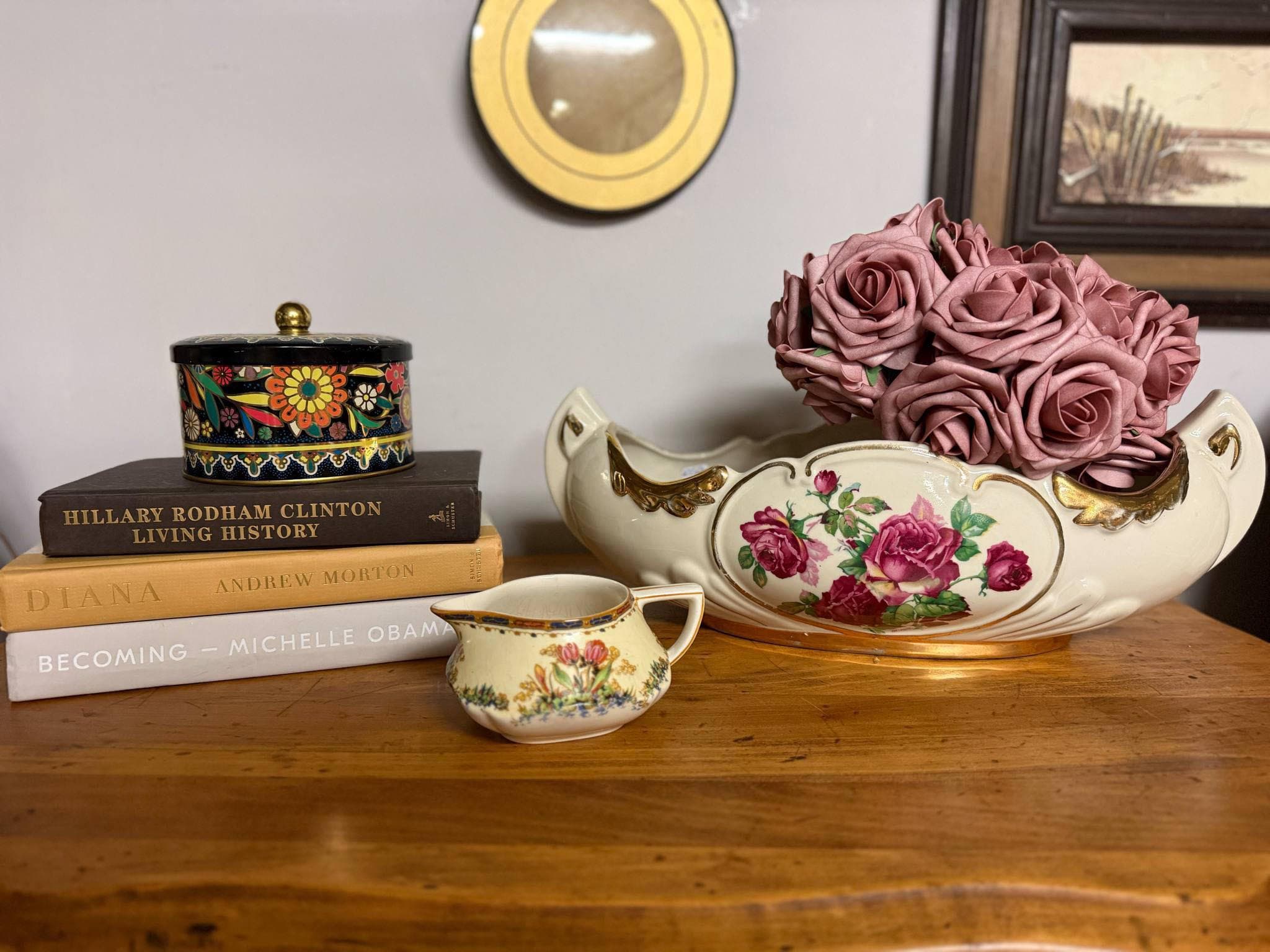 A tabletop vignette: a floral china bowl of mauve roses, a painted tin box, a cream pitcher, and a stack of hardcover books