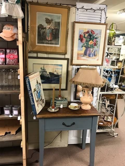 A blue-grey painted side table with a fringed vintage lamp, framed prints, and collectibles in a styled booth
